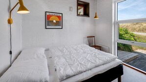 Ein Schlafzimmer mit Holzboden, einem Bett mit weißer Bettwäsche, zwei Wandleuchten, einem gerahmten Bauhaus-Poster und einem Fenster mit Blick auf Grünflächen.