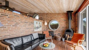 Eine gemütliche Terrasse mit Backsteinmauer, einem schwarzen Ledersofa, einem runden Tisch mit Obst und einem Kaminofen.