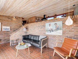 A cozy living area with a brick wall, a black leather sofa, a wooden chair, a small round table with a fruit bowl, and a painting of two people.