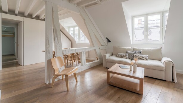 A loft-style living room with a high ceiling, exposed beams, a white sofa, a wooden coffee table, and a dining area with a white table and chairs.
