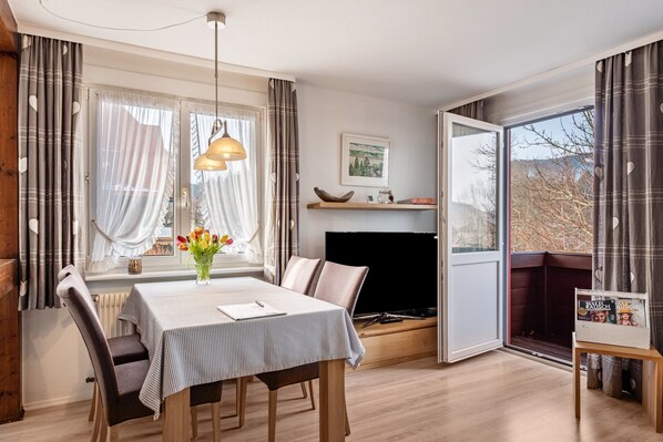 A dining area with a table, chairs, a vase of flowers, a television, and a view through an open door.
