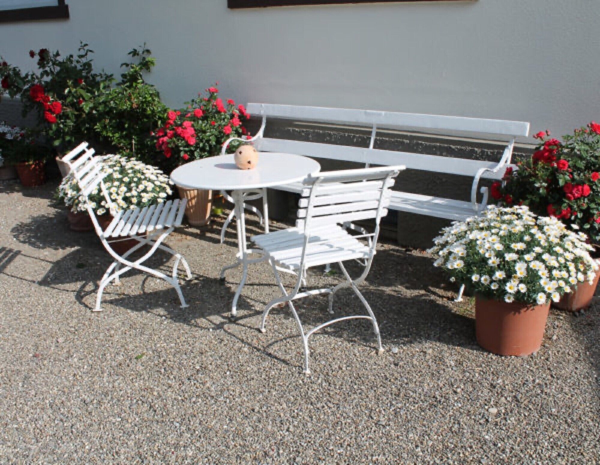 Ein weißer Außenbereich mit Sitzgelegenheit, einem runden Tisch und zwei Stühlen, umgeben von Kübelpflanzen mit roten und weißen Blüten.