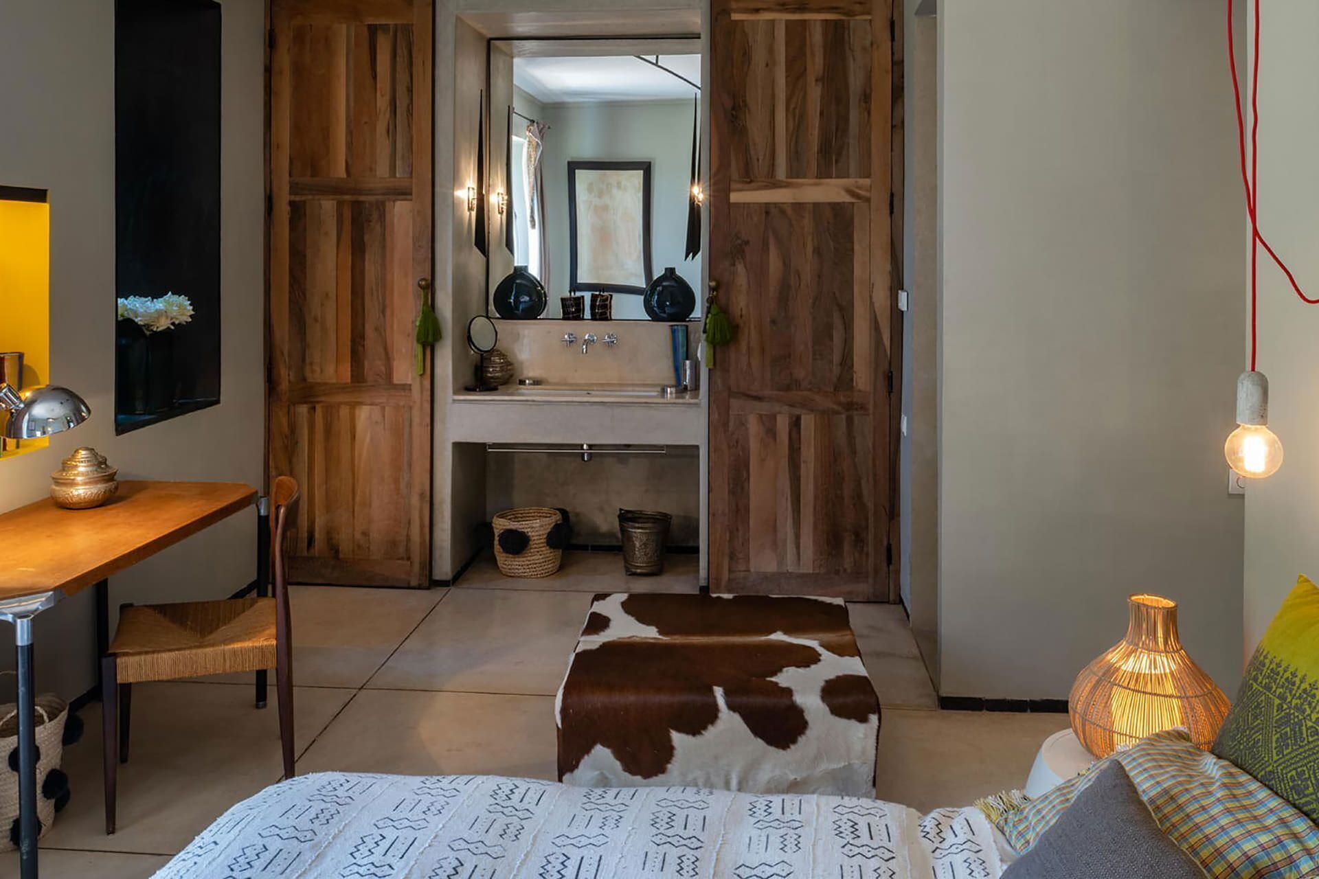 A room with a wooden wardrobe, a desk with a lamp, a bed with patterned bedding, and a bedside table with a vase.