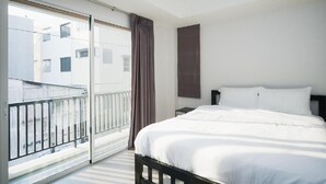 Habitación de hotel con una cama grande, una mesita de noche, una ventana con vistas a edificios y un balcón con barandilla.