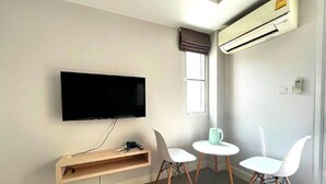 Una habitación moderna con televisión de pantalla plana en la pared, dos sillas blancas alrededor de una mesita redonda y un equipo de aire acondicionado en la pared.