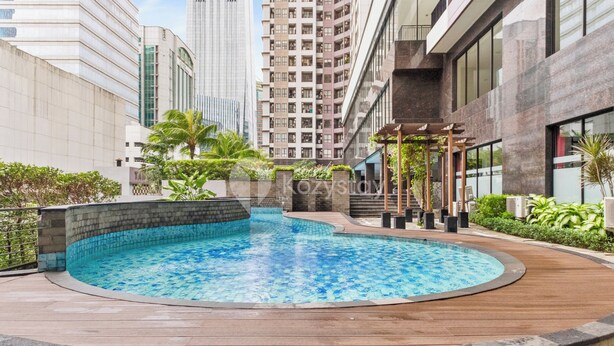 A swimming pool with a wooden deck, surrounded by greenery and modern buildings.