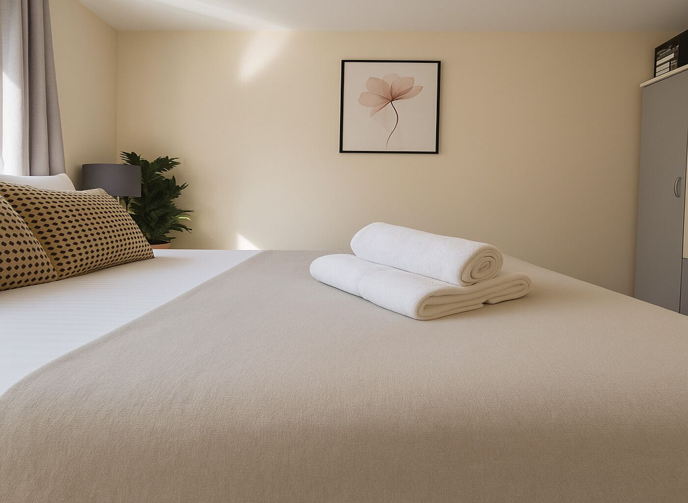 A neatly made bed with a rolled white towel, a framed flower artwork on the wall, and a plant beside the bed.