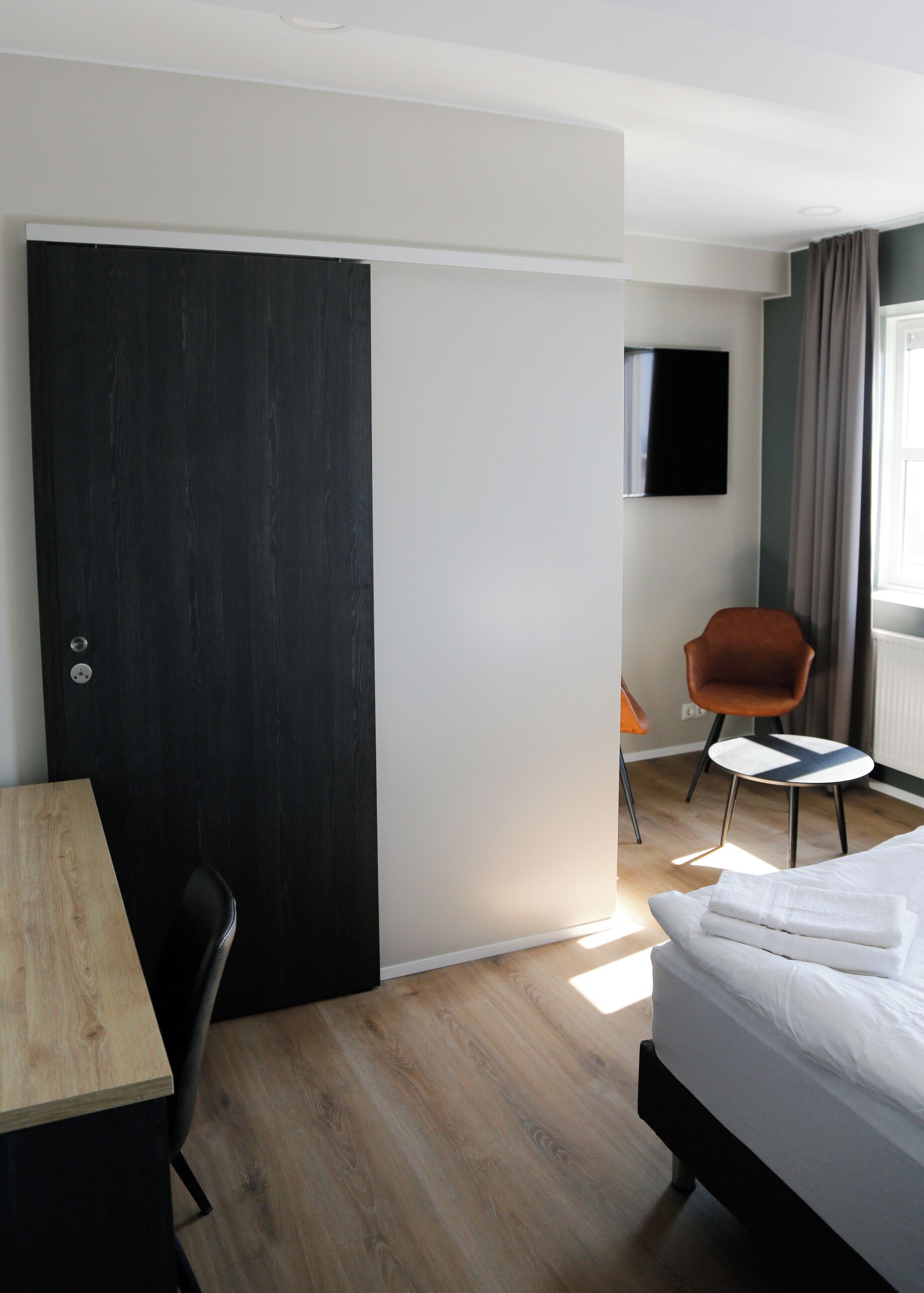 Ein Hotelzimmer mit einem Bett, einem Schreibtisch, einem Stuhl, einem an der Wand befestigten Fernseher und einem Fenster mit Vorhängen.