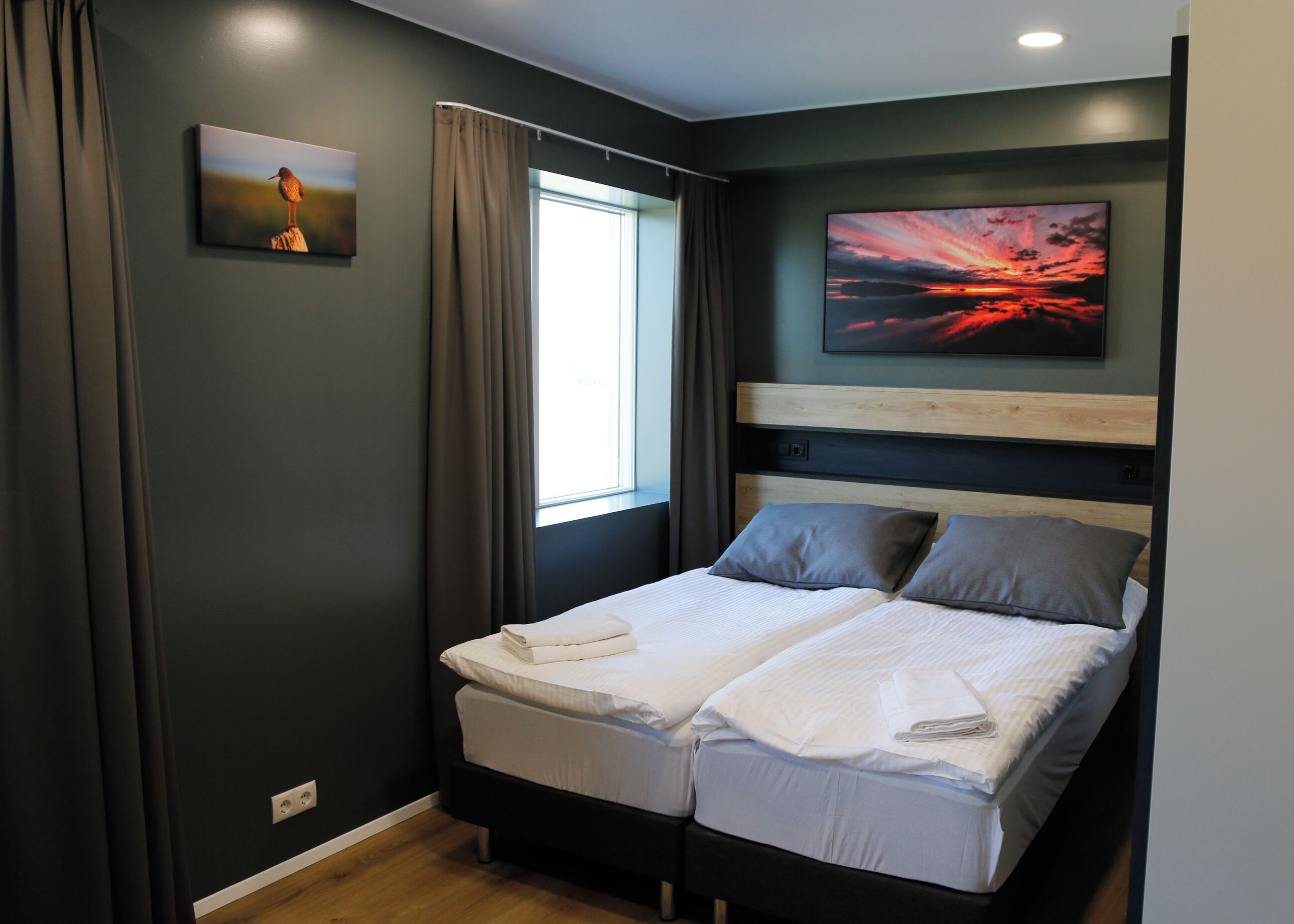 A double bed with a wooden headboard, grey bedding, and a window with curtains.