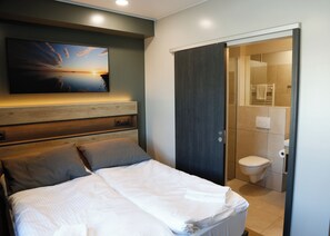 A bedroom with a bed, a headboard, a wall-mounted TV, and a door leading to a bathroom. - Gistihúsið Hofsósi (Hofsós)