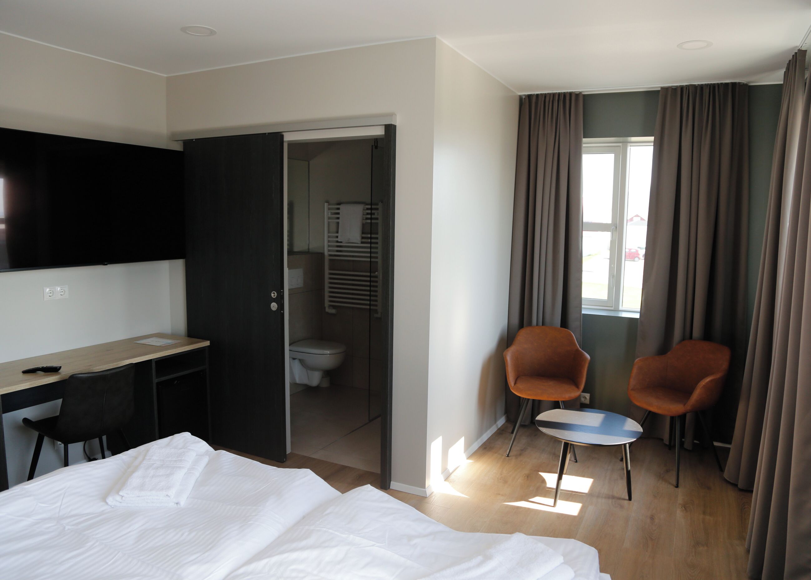 Habitación de hotel con cama, escritorio, dos sillas, televisor y baño a la vista a través de una puerta abierta.