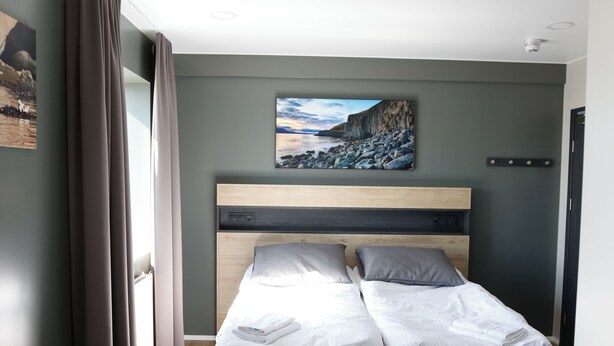 A room with two single beds, a headboard with control buttons, a painting of a coastal scene, and a window with curtains.