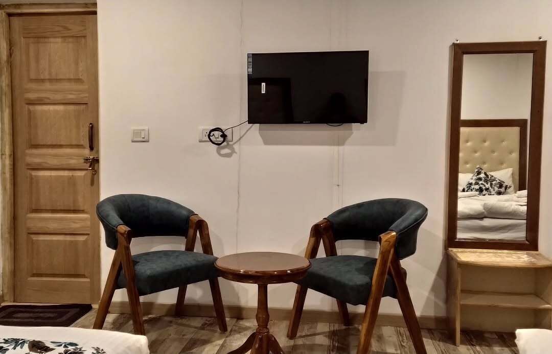 A room with a wooden door, a TV mounted on the wall, two upholstered chairs around a small wooden table, and a bed with a headboard and a mirror.