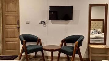 A room with a wooden door, a TV mounted on the wall, two upholstered chairs around a small wooden table, and a bed with a headboard and a mirror.