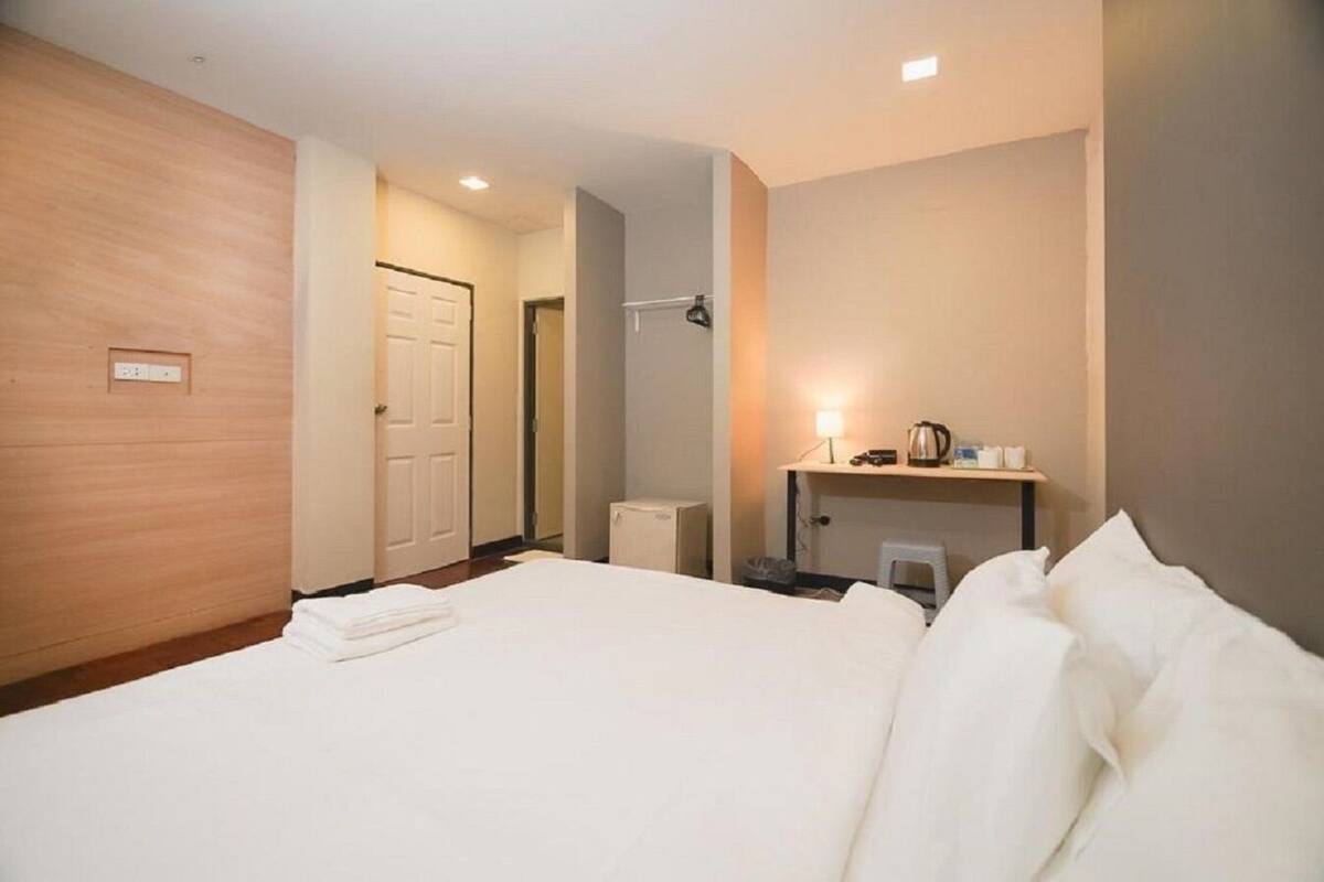 Habitación de hotel con una cama grande, un escritorio con lámpara y un taburete pequeño. Se ve una puerta que da a otra habitación y una lámpara en el techo.