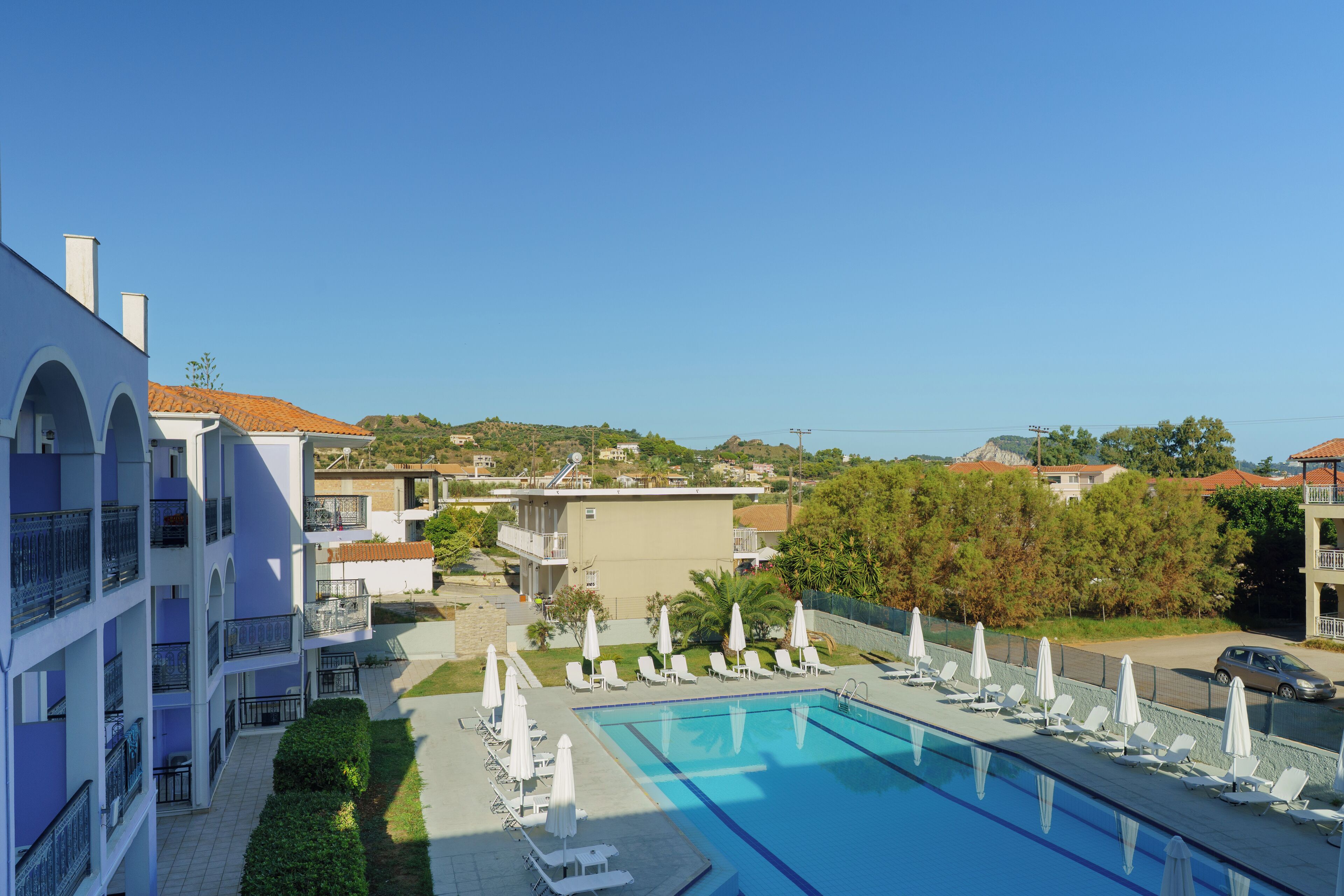 A pool area with lounge chairs and umbrellas, surrounded by buildings with balconies and a clear sky.