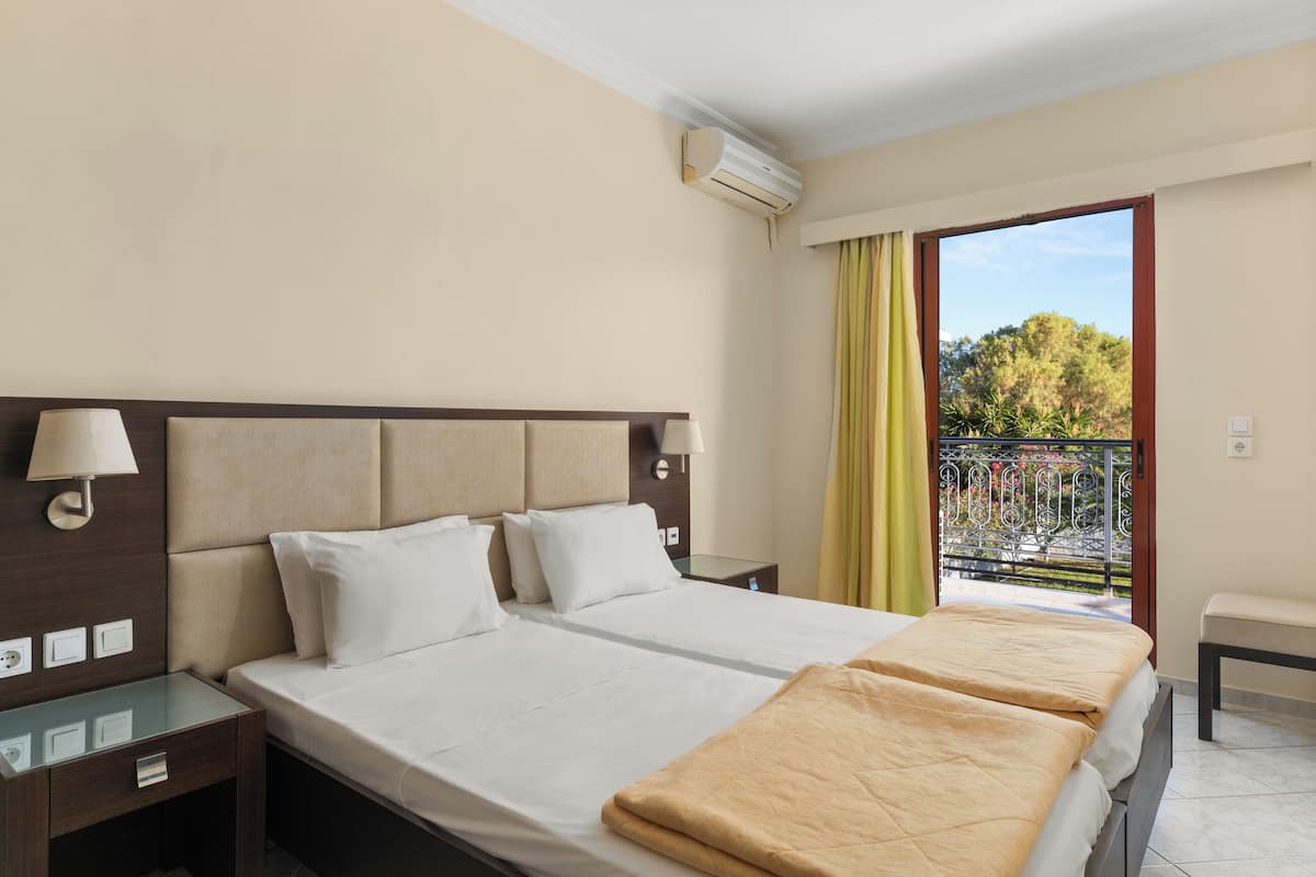 A hotel room with a large bed, bedside tables, a lamp, an air conditioner, and a balcony with a view of trees.