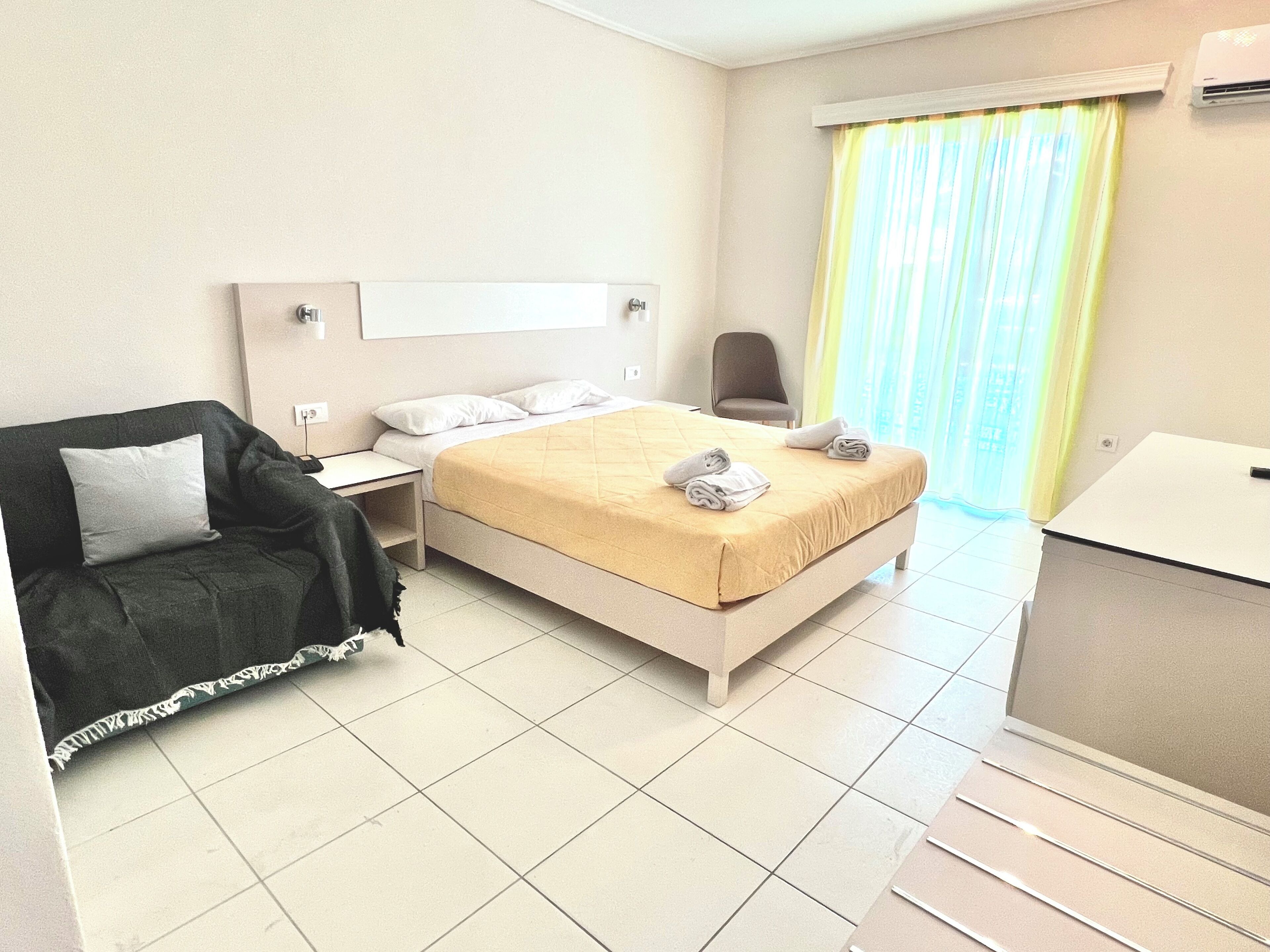 Quarto de hotel com cama, uma cadeira, escrivaninha e janela com cortinas.