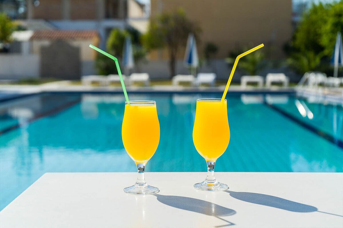 Two glasses of orange juice with straws on a white table by a swimming pool.