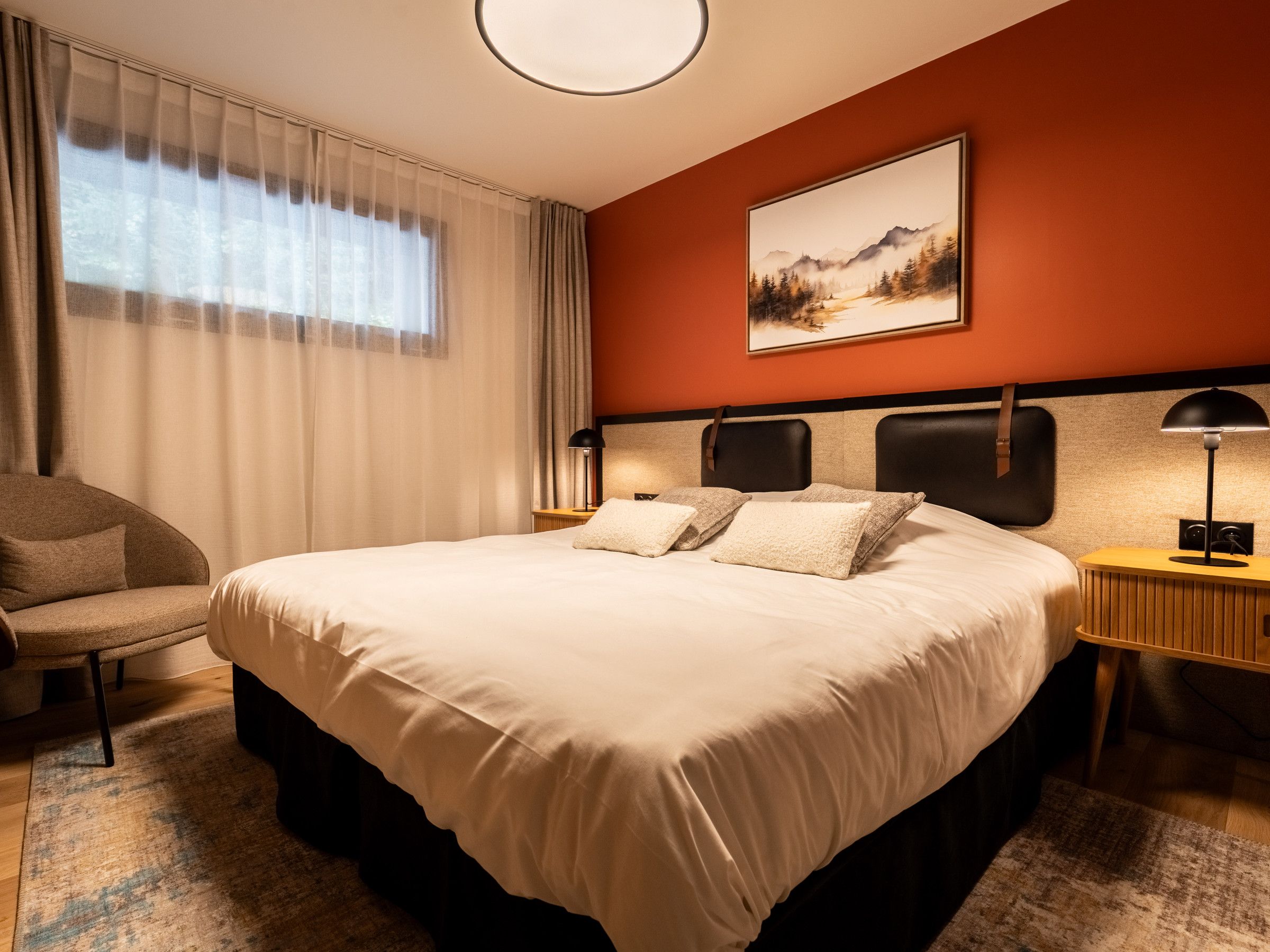A hotel room with a large bed, a chair, a bedside table with a lamp, and a painting on the wall.