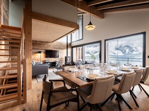 A modern dining area with a wooden table set for six, large windows offering mountain views, and a cozy seating area with a sofa and TV. - New Chalet in La Plagne - 3Lv, 12p, Sauna, Parking, WiFi (La Plagne Montalbert)