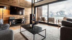 A modern living room with a large TV, wooden walls, a fireplace, and a balcony with a mountain view.