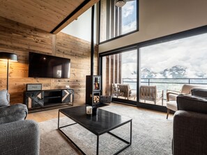 Una sala de estar moderna con un televisor grande, paredes de madera, chimenea y un balcón con vista a la montaña.