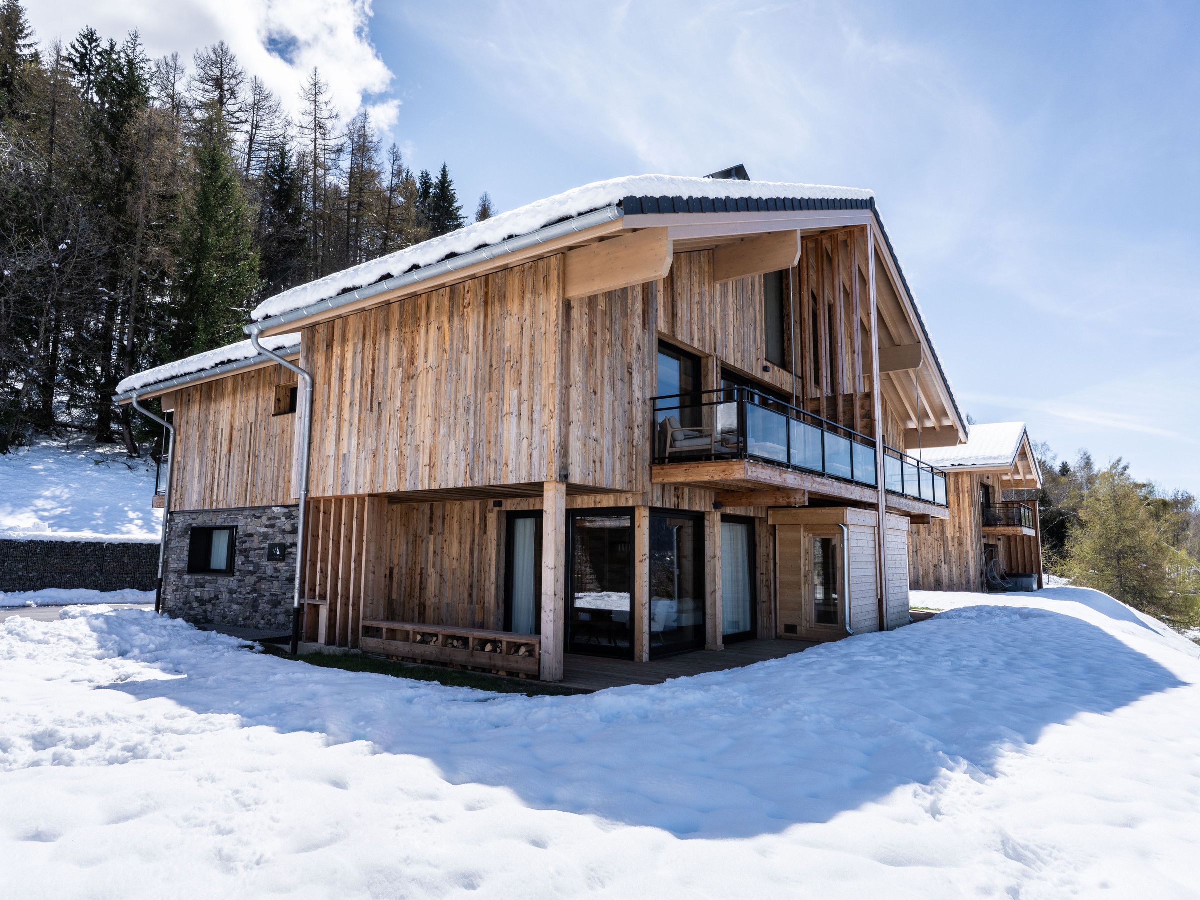 A wooden chalet with a balcony, stone foundation, and a snowy landscape.