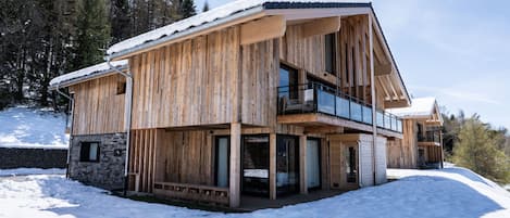 A wooden chalet with a balcony, stone foundation, and a snowy landscape.