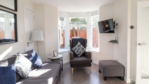 A modern living room with a sofa, armchair, and ottoman, a small side table, a TV mounted on the wall, and large windows with a view of the outside. - View Cottage (Inverness)
