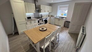 A compact kitchen with white cabinetry, a wooden dining table, and a window providing natural light. - 1 Links Place (Nairn)