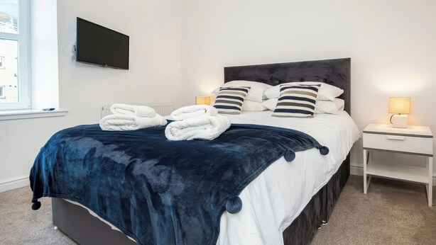 A neatly made bed with a dark blue blanket and white linens, a bedside table with a lamp, and a TV mounted on the wall. - 4 Lower Kessock Street (Inverness)