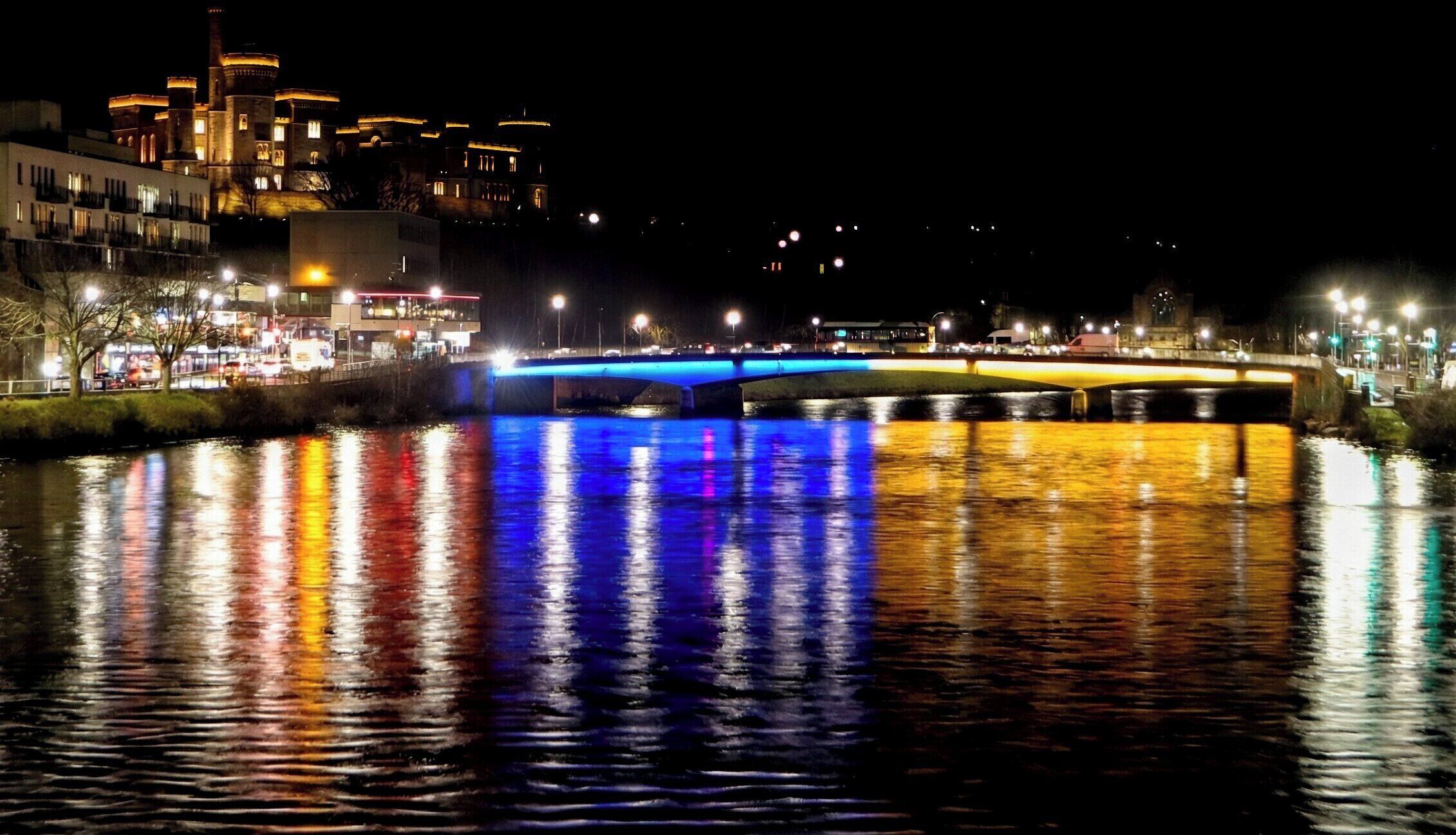 A nighttime view of a river with a bridge reflecting colorful lights, a castle-like building in the background, and a cityscape with illuminated buildings.