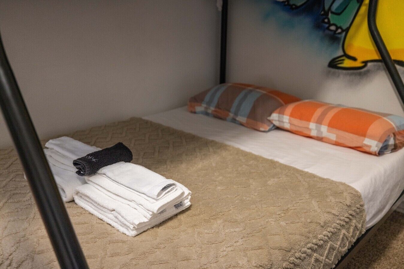Una cama litera con una toalla blanca doblada en la parte inferior, una toalla negra en la misma, una colcha color beige en la parte superior y dos almohadas estampadas en cuadros.
