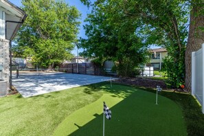 Un jardin à l’arrière d’une maison, comprenant un court de tennis, une pelouse artificielle, des arbres et une clôture.