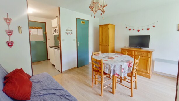 Una sala de estar acogedora con sofá, una mesa de comedor con sillas, televisión y una cocina visible a través de un ventanal.