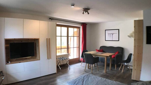 A compact living space with a sofa, a dining table with chairs, a TV mounted on a wall, and a window with curtains. - Apartments "Laber" for 2 persons, with kitchen (Oberammergau)