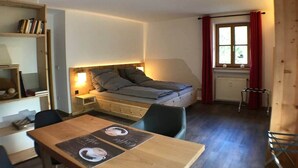 A compact room with a bed, a small table with a menu, a chair, a bookshelf, and a window with curtains. - Apartments "Laber" for 2 persons, with kitchen (Oberammergau)