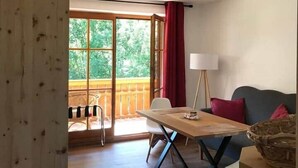 Et værelse med trægulv, et spisebord med stole, en sofa, en lampe og udsigt til grøn natur gennem store vinduer.