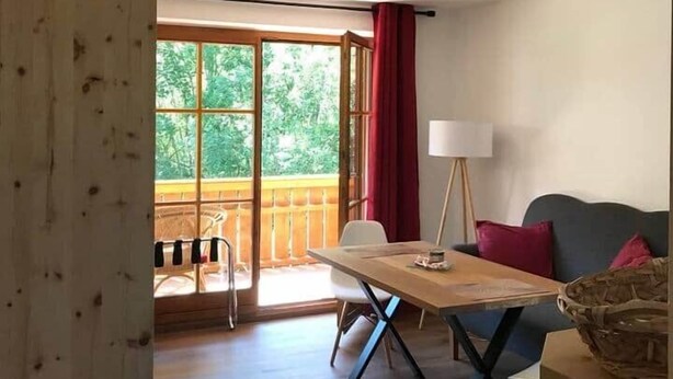 A room with a wooden floor, a dining table with chairs, a sofa, a lamp, and a view of greenery through large windows.