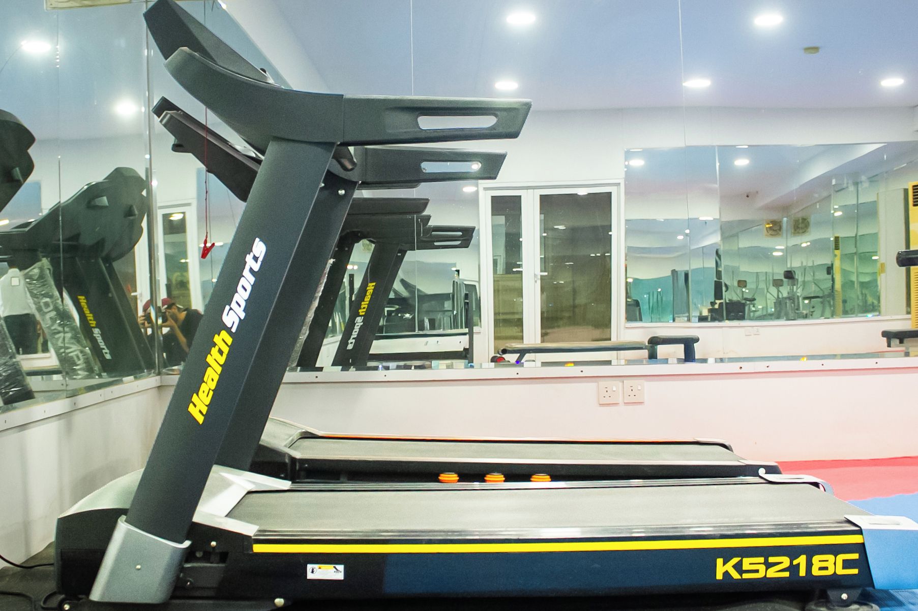 Una cinta de correr con pantalla digital y panel de control, ubicada en un gimnasio con paredes de espejo y equipamiento de ejercicio.