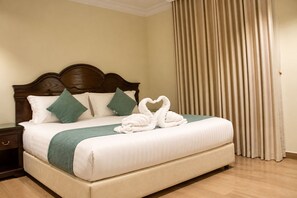 A hotel room with a large bed, a wooden headboard, a green throw blanket, and a swan-shaped towel arrangement. - Gulf Home (Al Khobar)