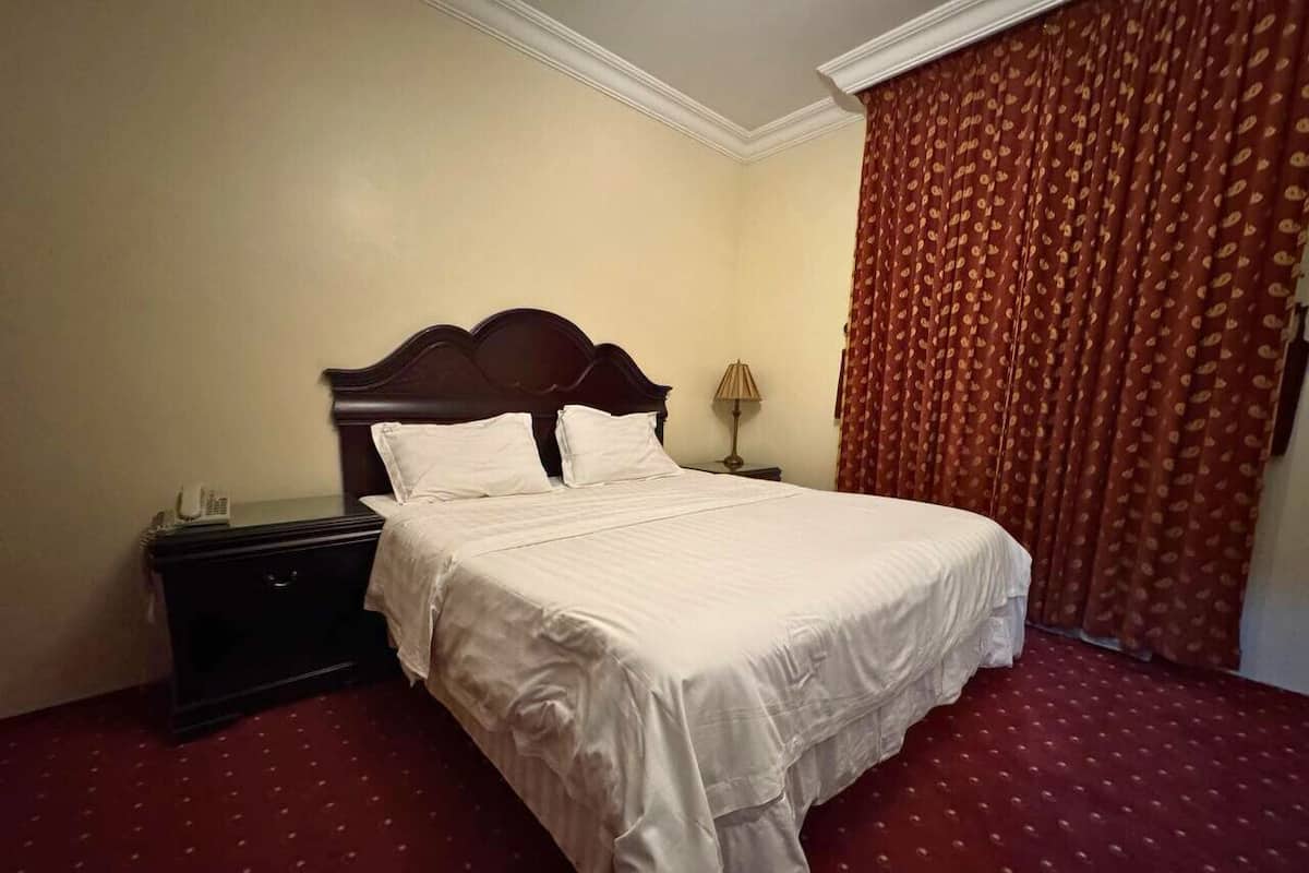 A hotel room with a large bed, a nightstand, a lamp, and a red patterned curtain.