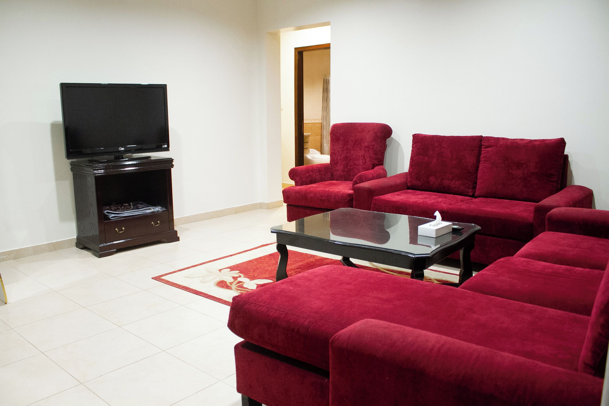 A living room with a flat-screen TV, a dark wooden entertainment unit, red velvet sofas, and a glass coffee table.