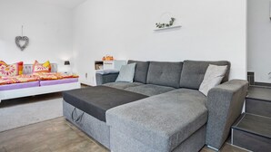 Un salon moderne avec un canapé d’angle gris, un canapé blanc agrémenté de coussins colorés, une petite table et un objet décoratif en forme de cœur accroché au mur.
