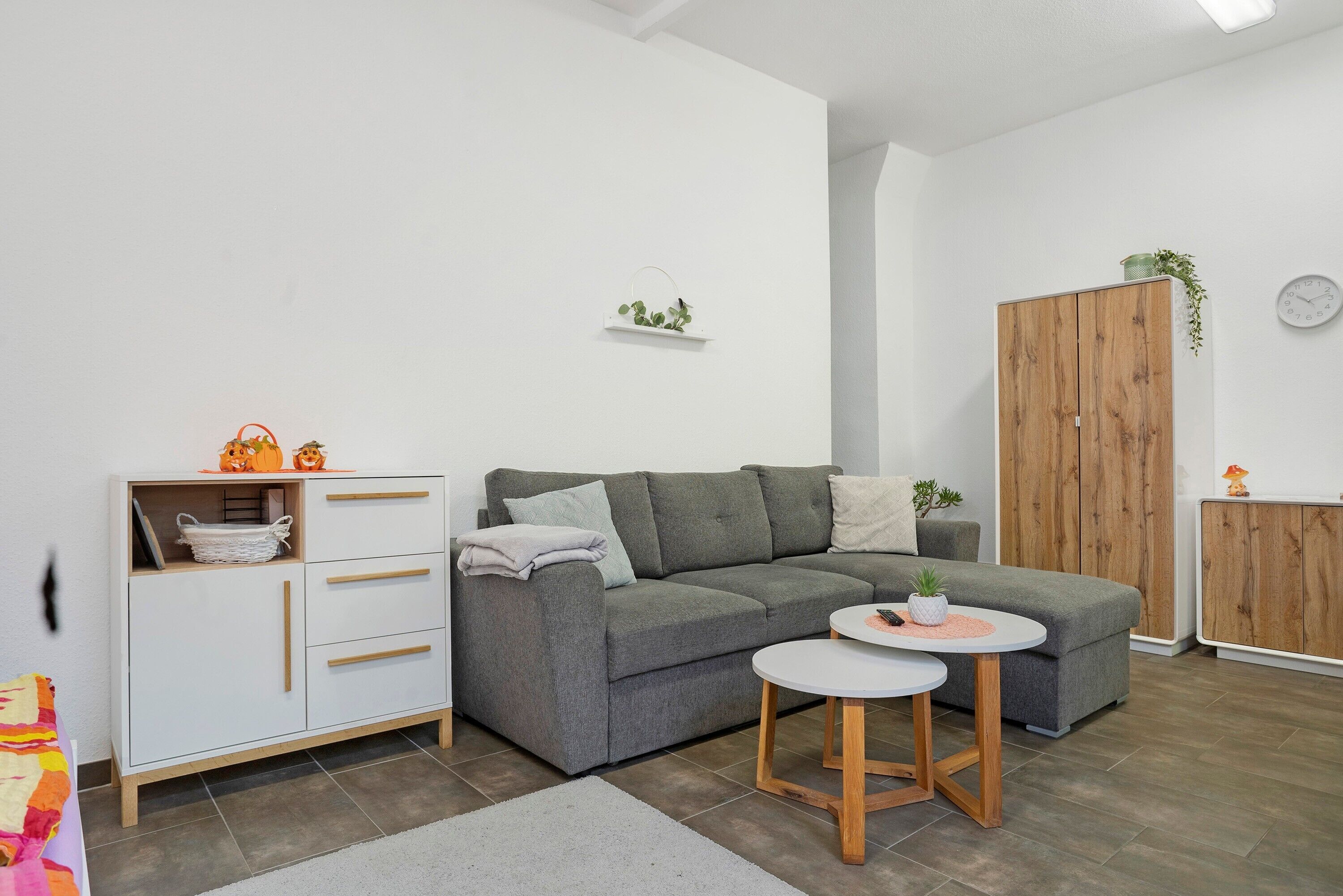 Een moderne woonkamer met een grijze hoekbank, een witte kast met lades, een klein rond tafeltje en een houten garderobe.