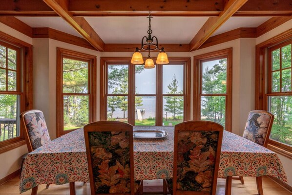 A dining area with a table set for four, large windows with a view of trees and a body of water, wooden beams on the ceiling, and a chandelier. - Whispering Pines Cabin on Devils Lake (Webster)