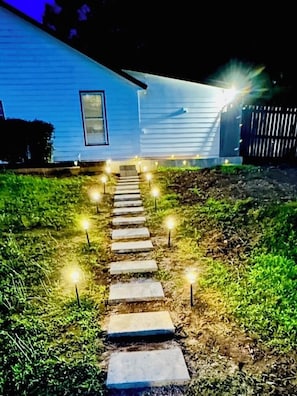 Un chemin menant à une maison dotée d’une fenêtre et d’une porte, entourée de pelouse et de marches en pierre éclairées.