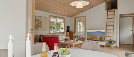 Ein modernes Wohnzimmer mit hoher Decke, Holzbalken, einem Esstisch mit Kerzen, Flaschen und einer Schüssel Trauben.
