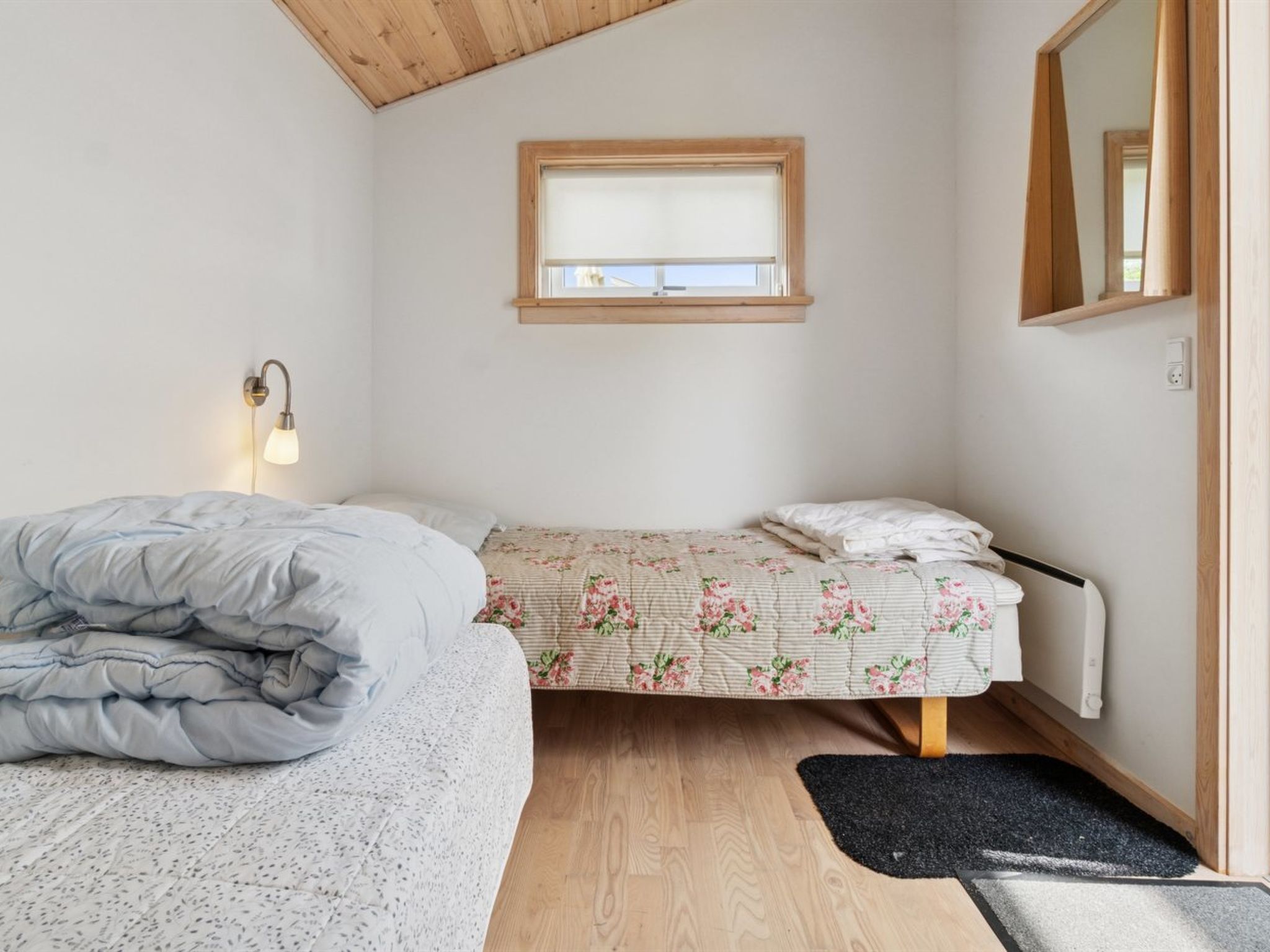 A bedroom with a bed, a bedside table with a lamp, a window, a radiator, and a mirror.